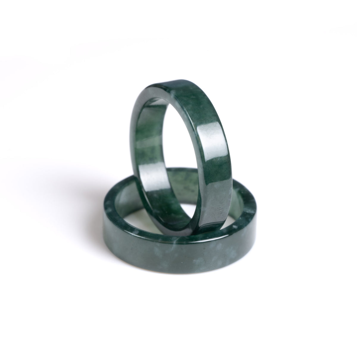 One natural jade ring positioned vertically in the middle of another natural jade ring that is laying flat on a white background. Both jade rings are in deep green colour. 